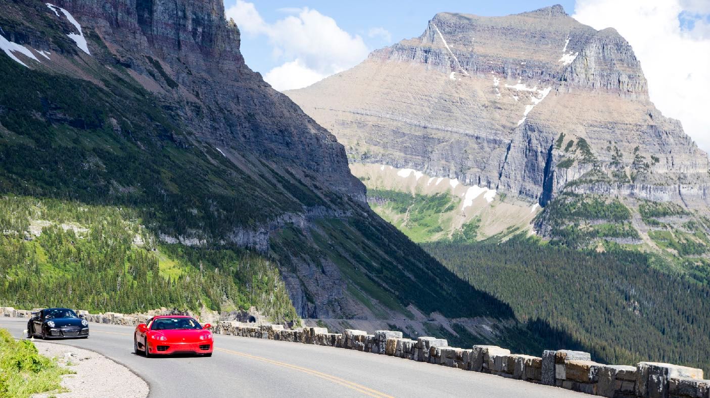 A red Ferrari and a black Porsche on Going-to-the-Sun Road in Glacier National Park, cliffs and snowfields behind