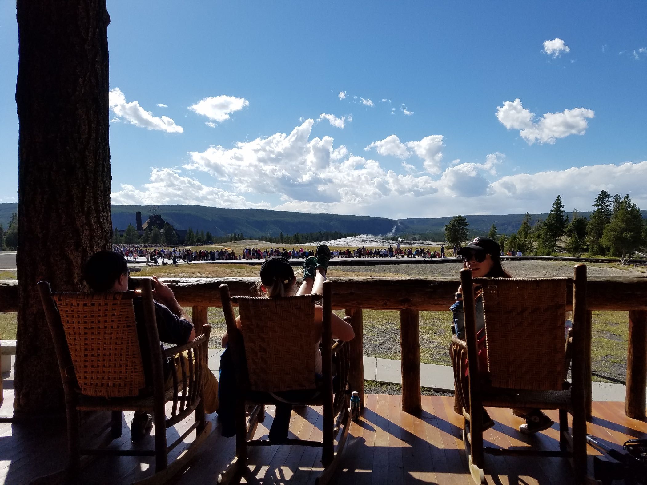 Guests in rocking chairs on a lodge porch, looking out over the Old Faithful geyser basin in Yellowstone